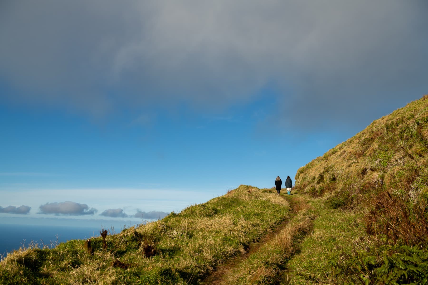 Two people walking a grassy ridge path high above the ocean